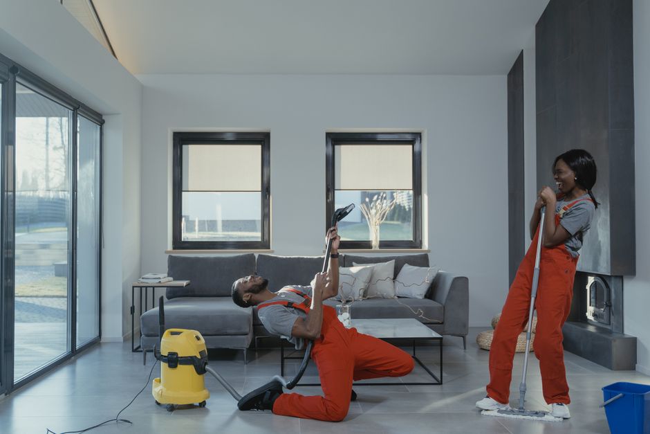 Fun and engaging cleaning session with a man and woman in overalls using cleaning tools in a modern living room.